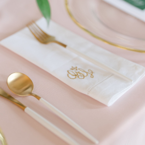 A white and gold embroidered wedding napkin with a monogram logo sat next to a plate with a fork resting on top